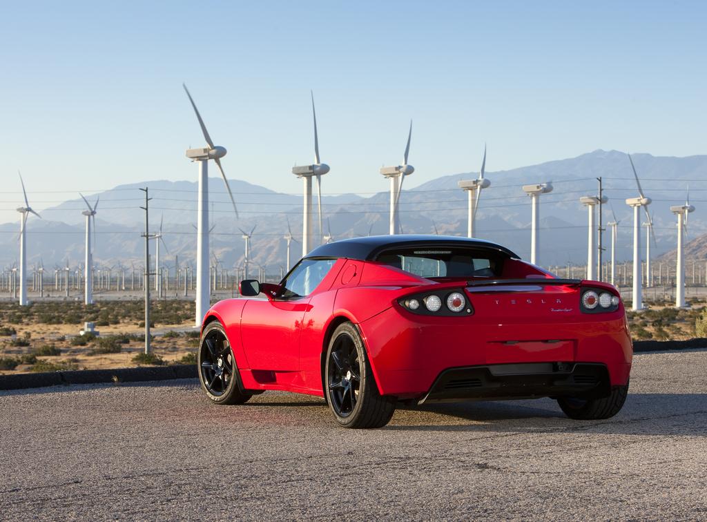 The back of a red Tesla Roadster in front of wind turbines